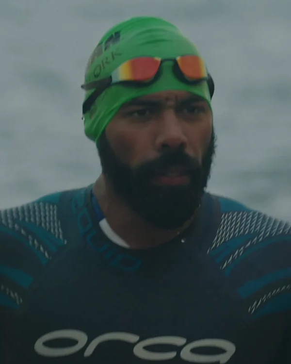 Leo in a wetsuit and green swim cap, looking straight at the camera with the sea behind him